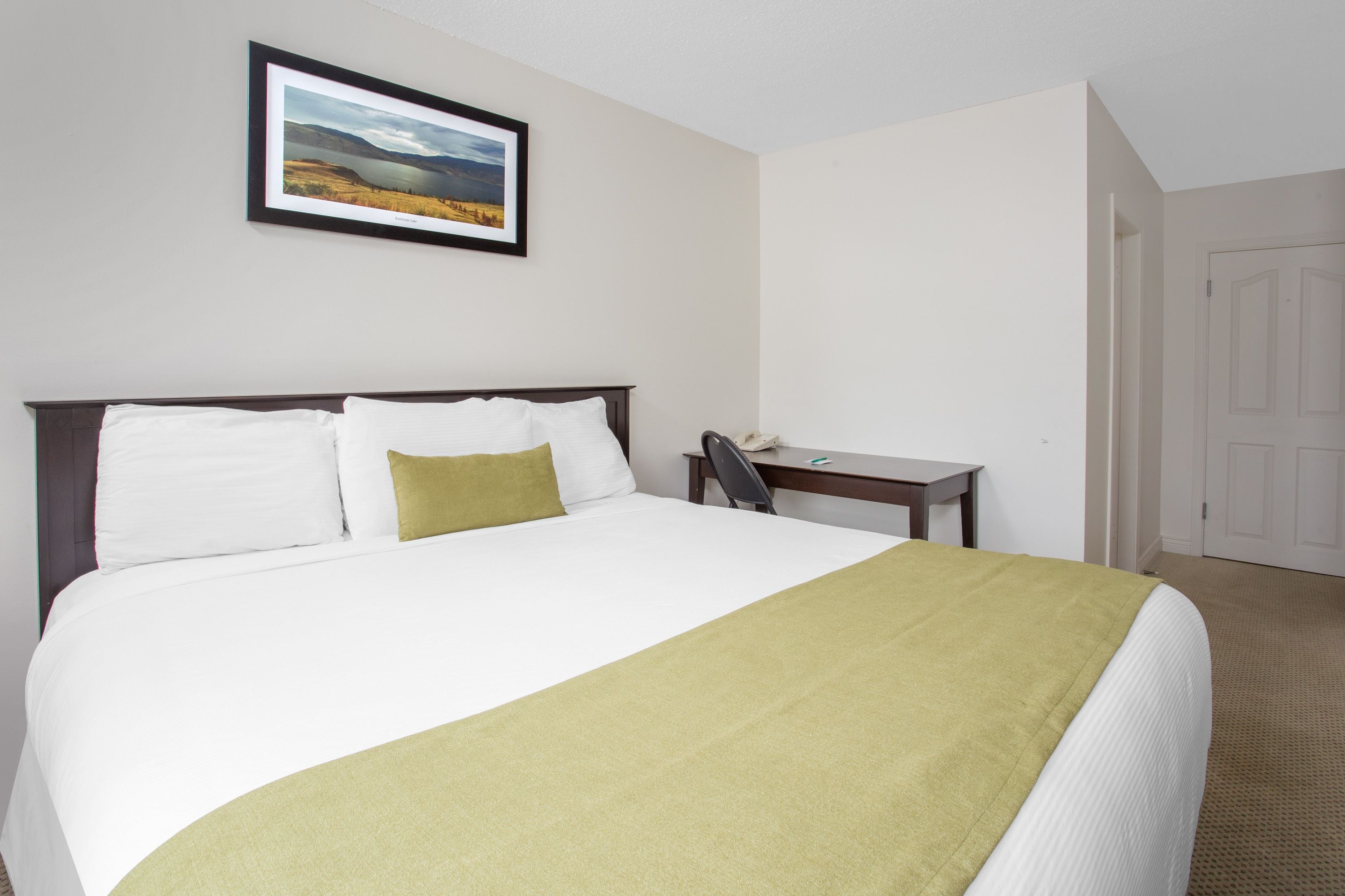 A neatly made bed with white and green linens, a framed landscape picture on the wall, and a small bedside table with a phone.