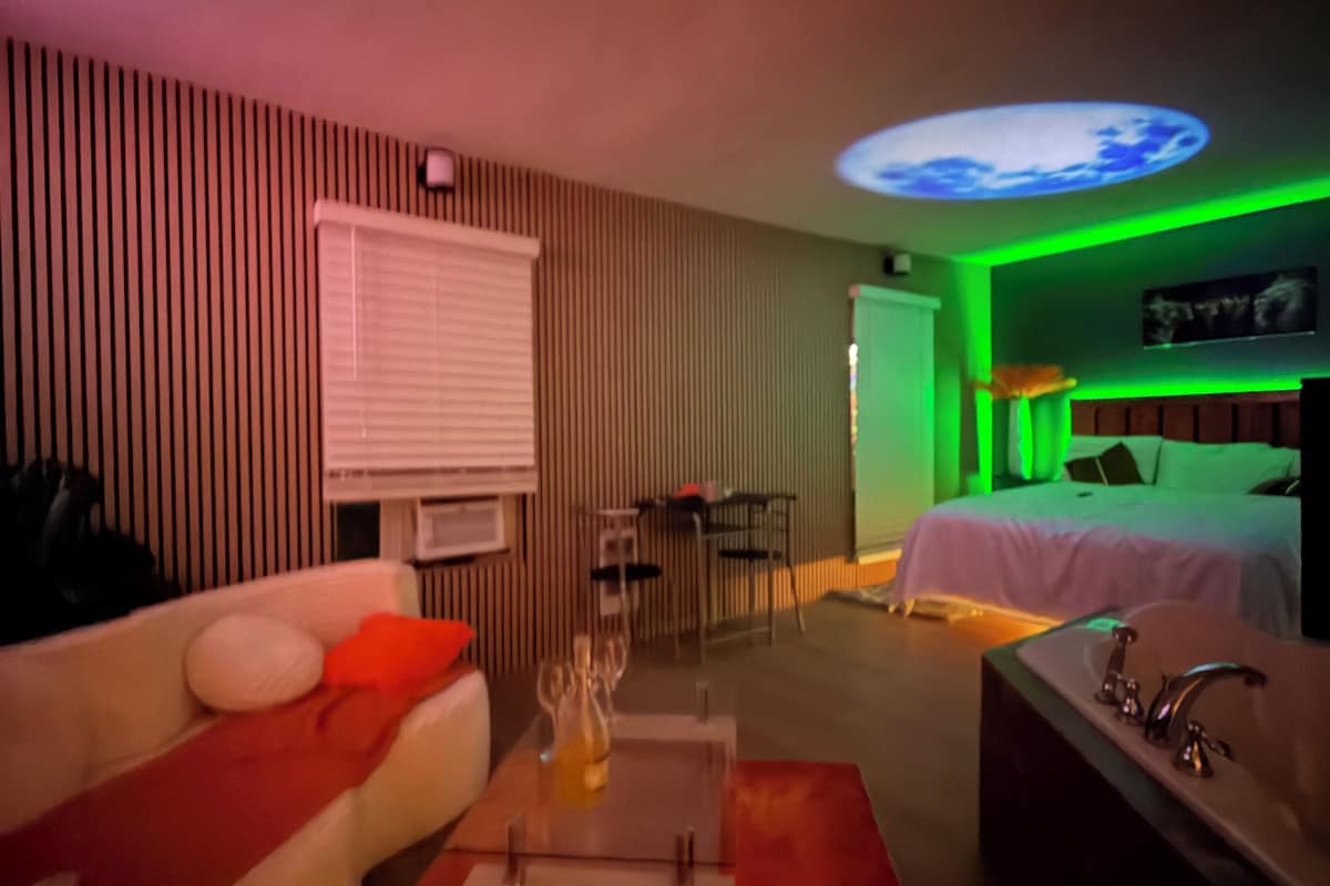 Habitació d'hotel moderna amb sofà, taula de cafè, llit, bany i lluminària al sostre.