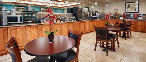 A hotel kitchen with wooden cabinets, a central island with food display, and a seating area with tables and chairs.