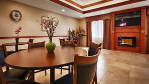A hotel dining room with a large wooden table, chairs, a green vase with flowers, a clock on the wall, and a fireplace.