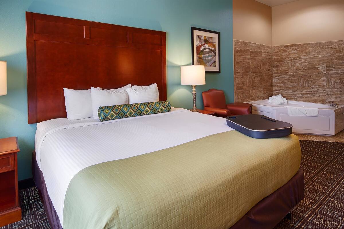 Habitación de hotel con una cama grande, dos lámparas de noche, una mesita de noche de madera, un sillón de cuero rojo, una pequeña mesa con una toalla y una pared con textura de piedra.