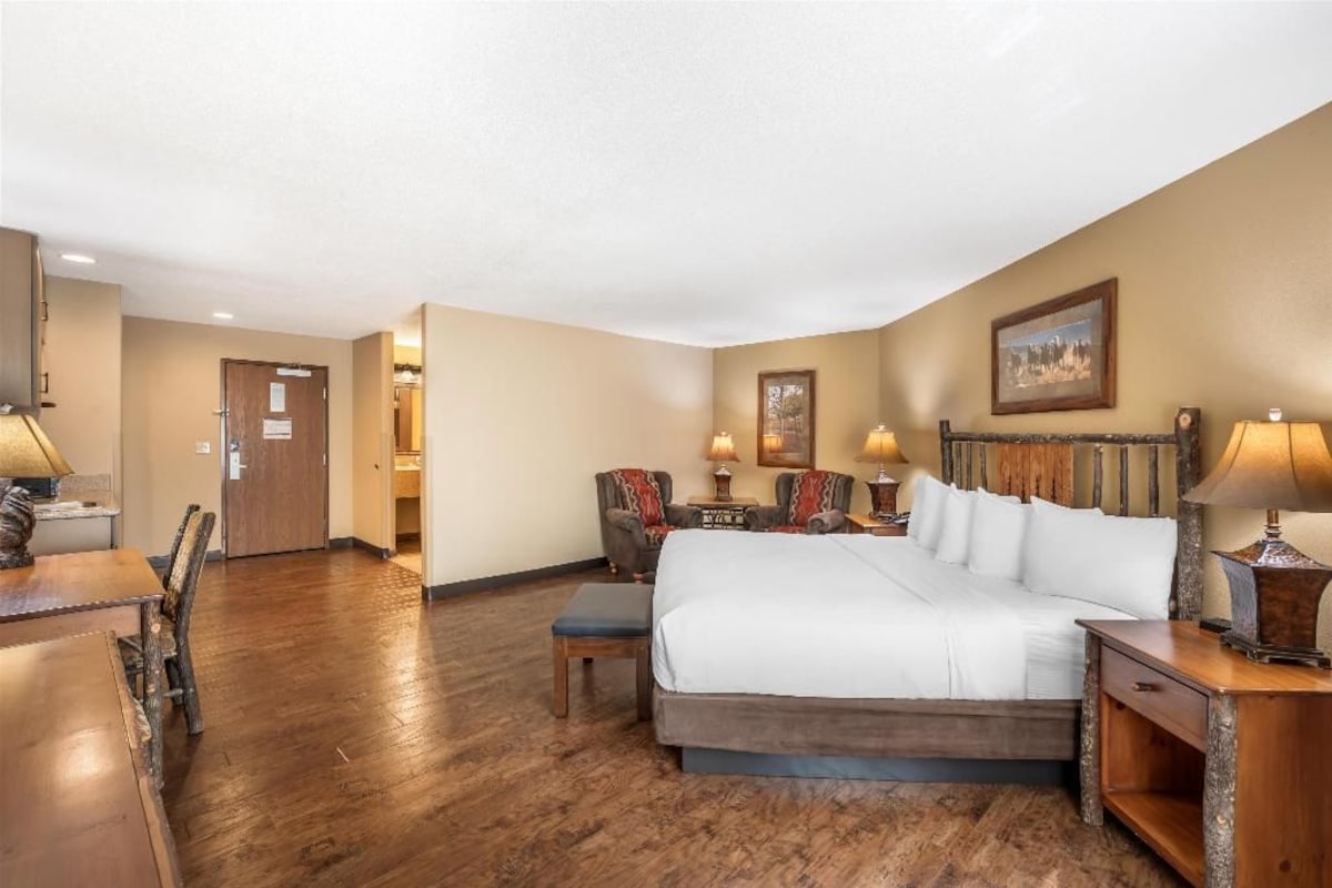 Habitación de hotel con una cama grande, dos mesitas de noche con lámparas, suelo de madera y una puerta que da a otra habitación.