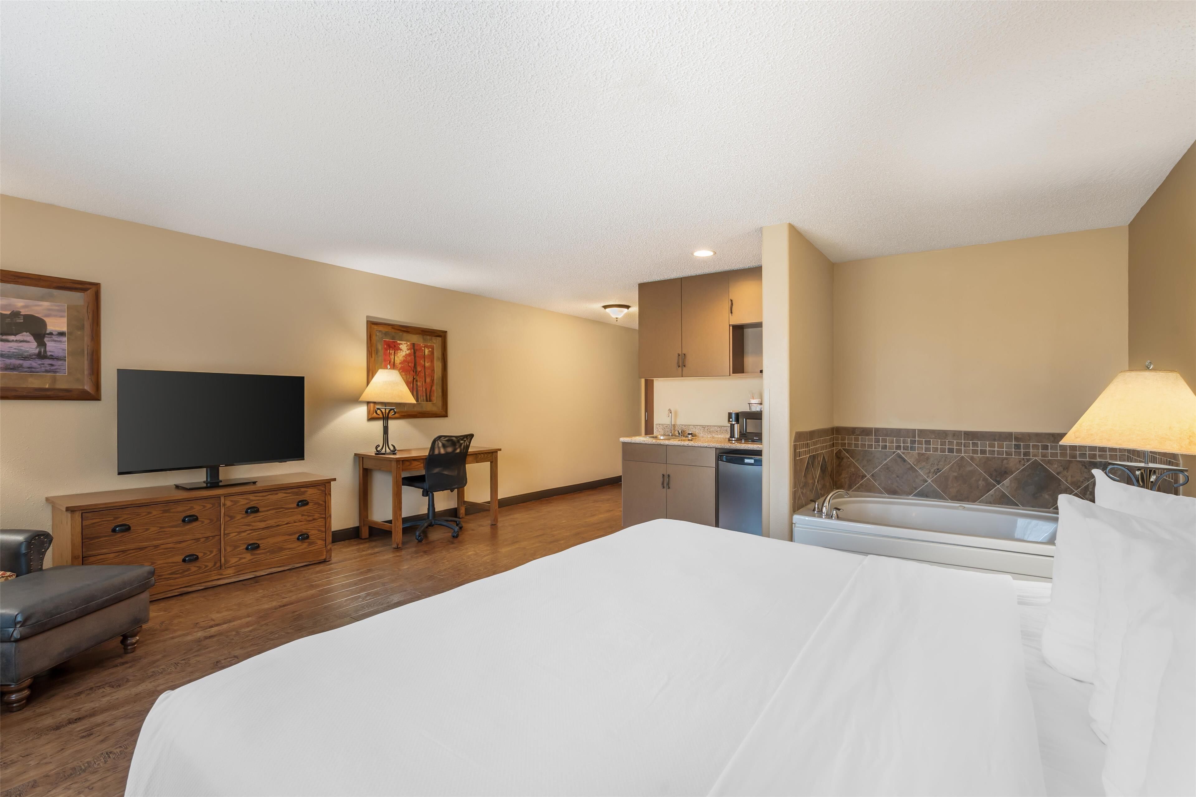 A hotel room with a bed, a desk, a television, a chair, a kitchen area, and a bathtub.