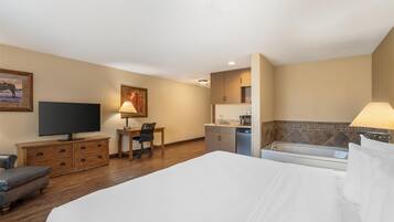 A hotel room with a bed, a desk, a television, a chair, a kitchen area, and a bathtub.