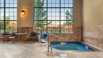 An indoor swimming pool area with a view of trees through large windows, a pool with a safety railing, and a seating area with chairs and a table.