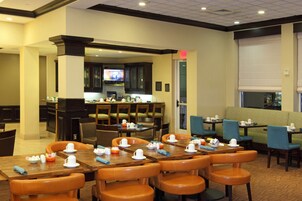 A hotel dining area with multiple tables and chairs, a kitchen in the background, and a television screen.