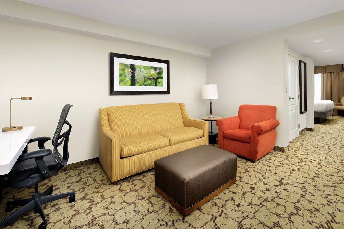 A hotel room with a yellow sofa, an orange armchair, a brown ottoman, a desk with a chair, and a framed picture on the wall.