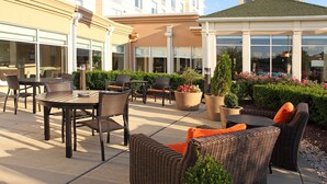 An outdoor patio with wicker furniture, potted plants, and a building in the background. - Hilton Garden Inn Frederick (Frederick)