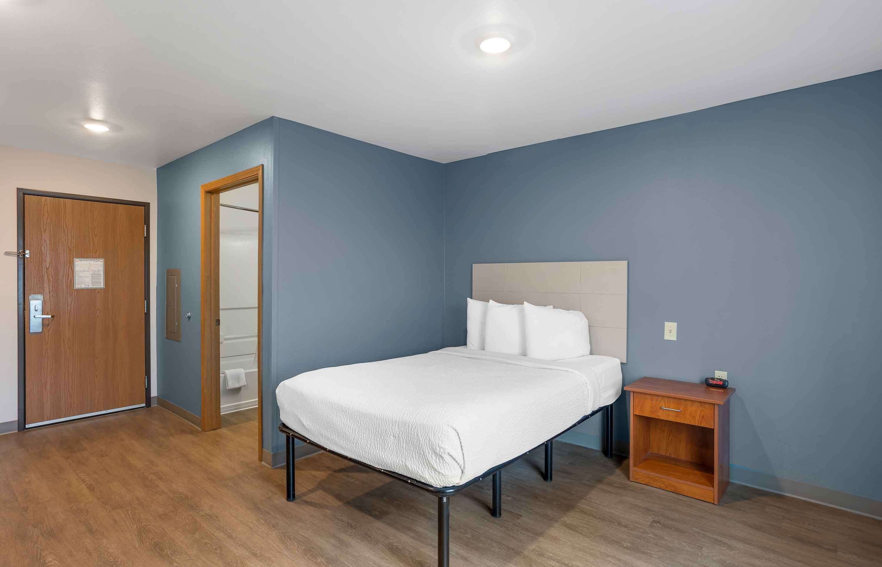 Un dormitorio con una cama, una mesita de noche, una puerta de madera y un baño a la vista a través de una puerta abierta.