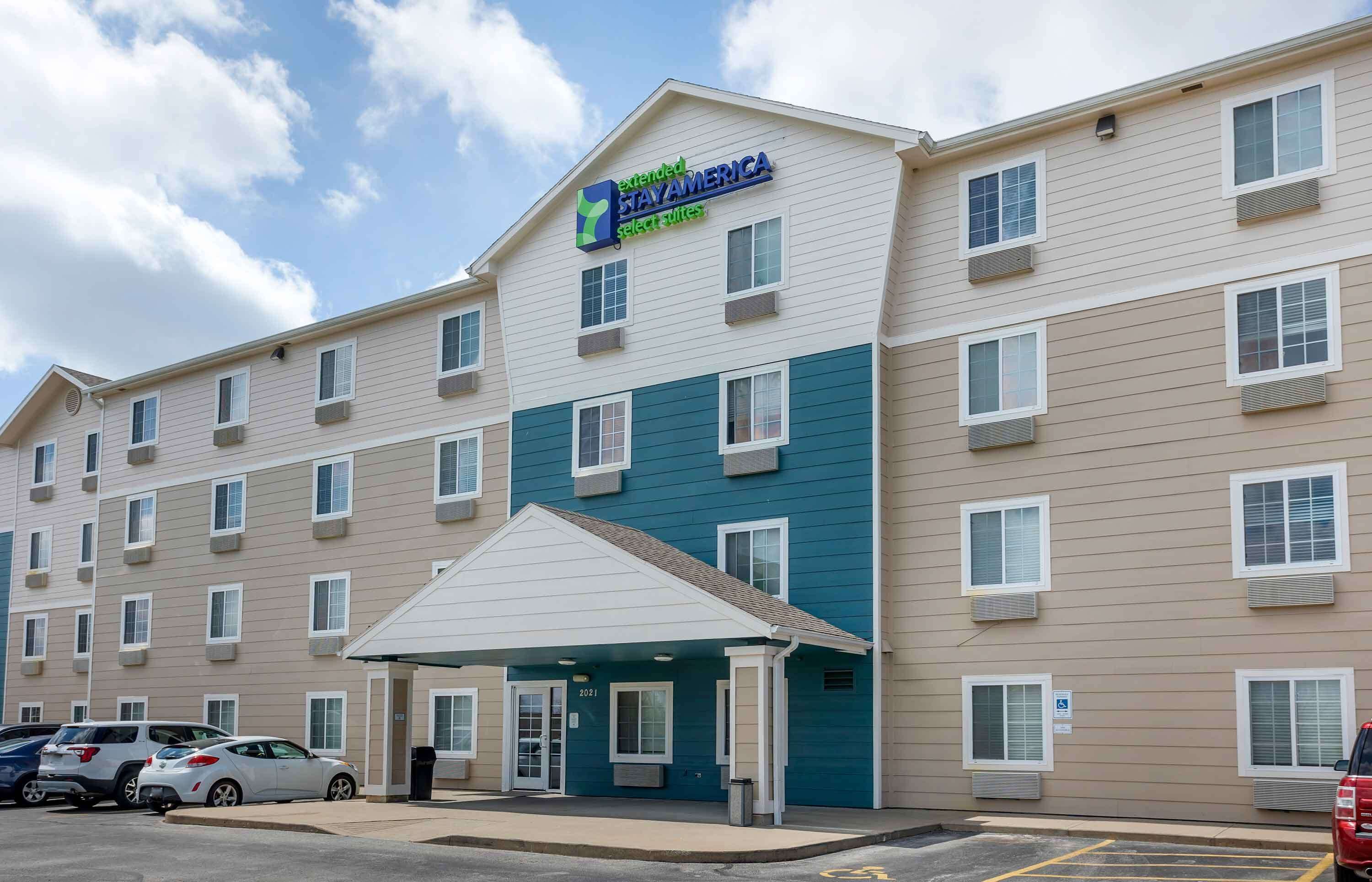 Il s’agit d’un hôtel dont l’enseigne indique « Extended Stay America Select Suites ». Il se distingue par un bâtiment à plusieurs étages doté d’un toit plat, d’une entrée abritée et d’un parking où sont stationnées plusieurs voitures.