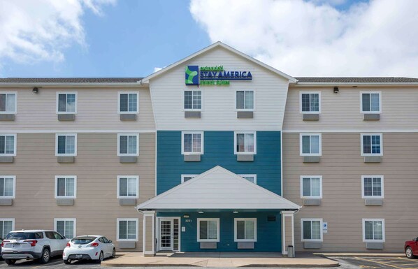 A multi-story hotel with a sign reading "StayAmerica." It features a central entrance with a covered area, multiple windows on each floor, and a parking lot with several cars.