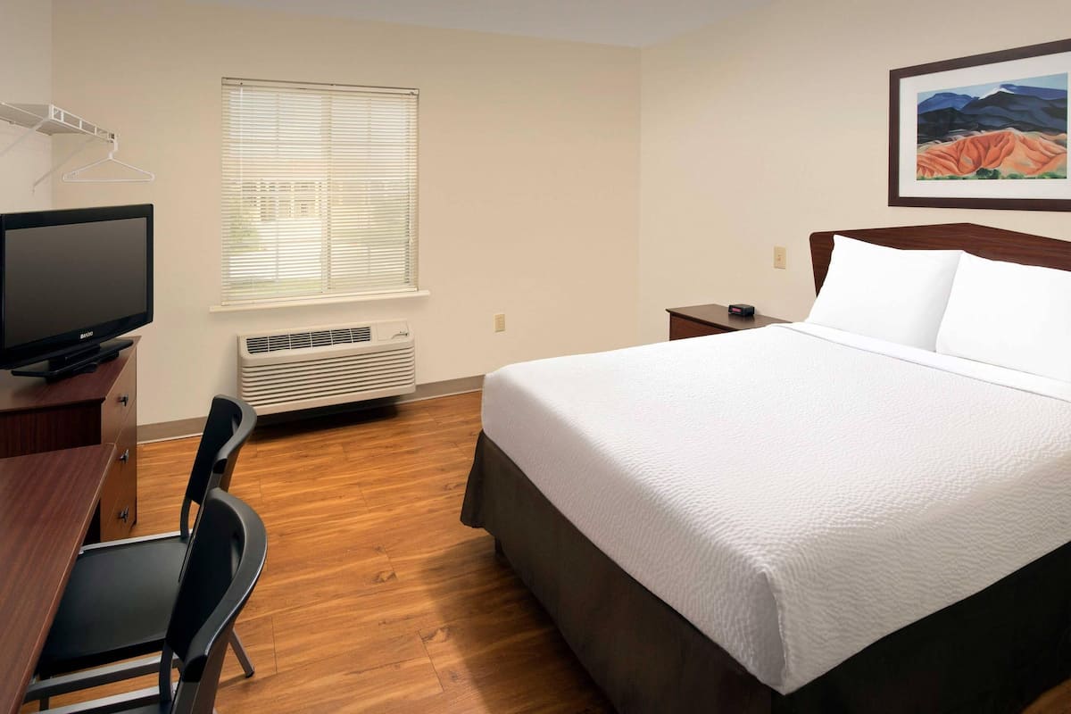 A hotel room with a bed, a desk, a chair, a television, a window with blinds, an air conditioner, and a framed picture on the wall.