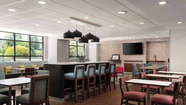 A modern restaurant with a bar area, seating with cushioned booths and tables, and a large window offering a view of greenery. - Hampton Inn Spring Lake Fayetteville (Spring Lake)