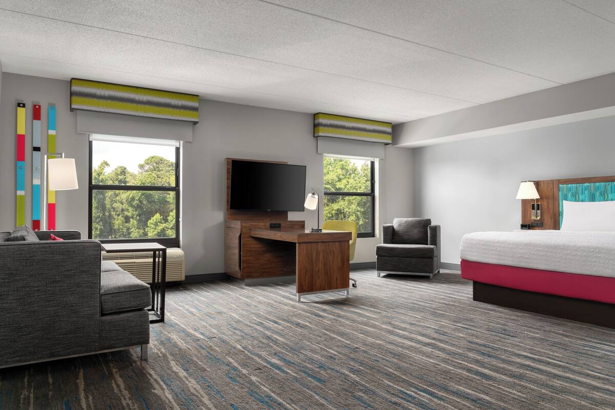 Ein Hotelzimmer mit Bett, Schreibtisch, Stuhl, Fernseher und großen Fenstern.