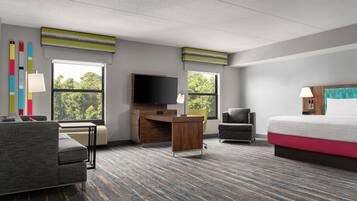 Una habitación de hotel con cama, escritorio, silla, televisor y ventanas amplias.