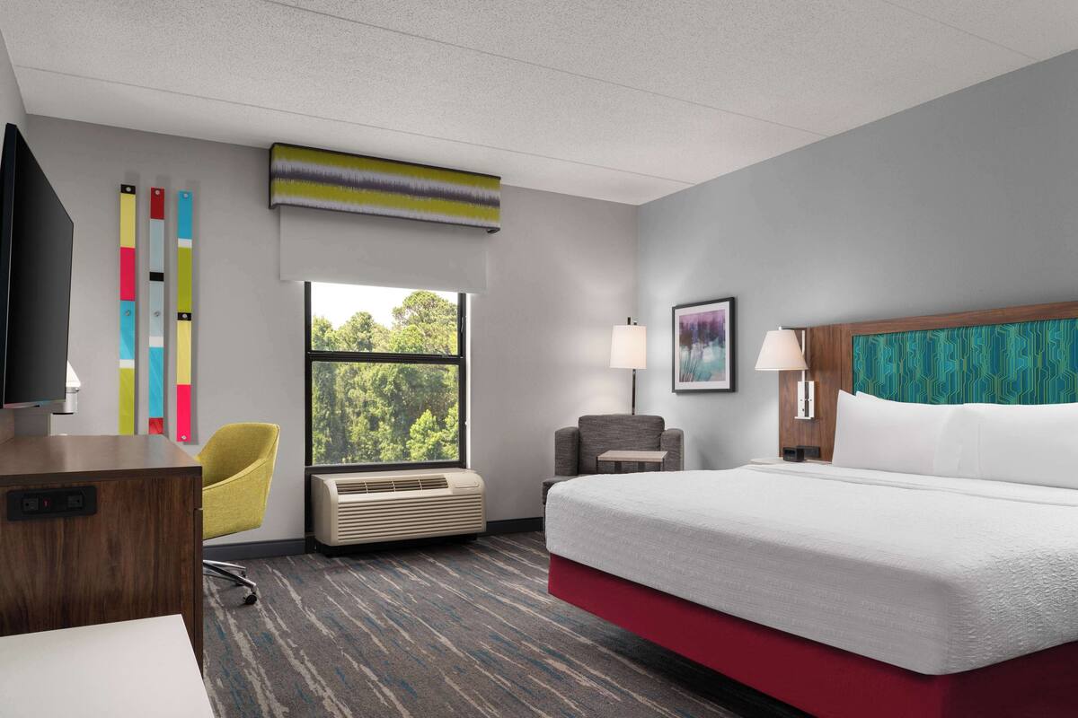 Ein Hotelzimmer mit einem großen Bett, einem Schreibtisch, einem Stuhl, einem Fenster mit Blick auf Bäume und Wandvorhängen mit bunten, senkrechten Streifen.