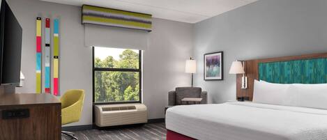 Une chambre d’hôtel avec un grand lit, un bureau, une chaise, une fenêtre donnant sur des arbres et un mur orné de stores verticaux aux couleurs vives.