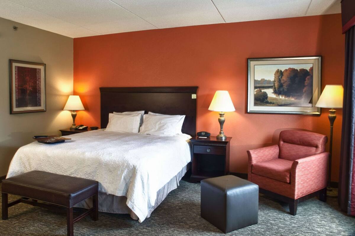 A hotel room with a bed, two bedside tables with lamps, a chair, a bench, and a framed picture on the wall.