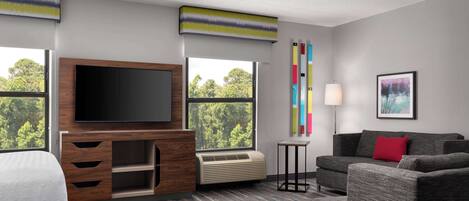 Habitación de hotel con una amplia televisión de pantalla plana, un sofá, una cama, una mesita y una ventana con vistas a árboles.
