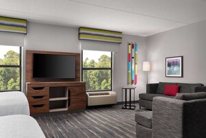 A hotel room with a large flat-screen TV, a sofa, a bed, a small table, and a window with a view of trees.