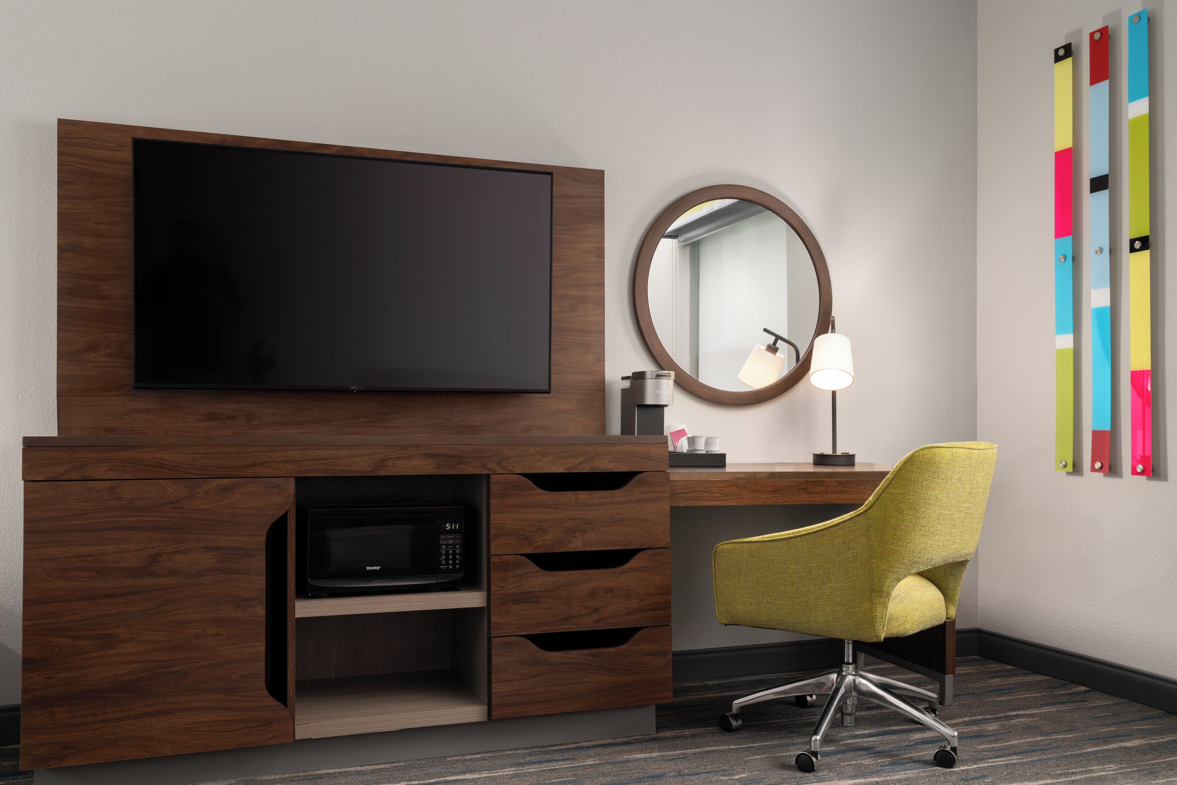 A hotel room with a flat-screen TV, a microwave, a desk with a chair, a round mirror, and a wall with colorful artwork.
