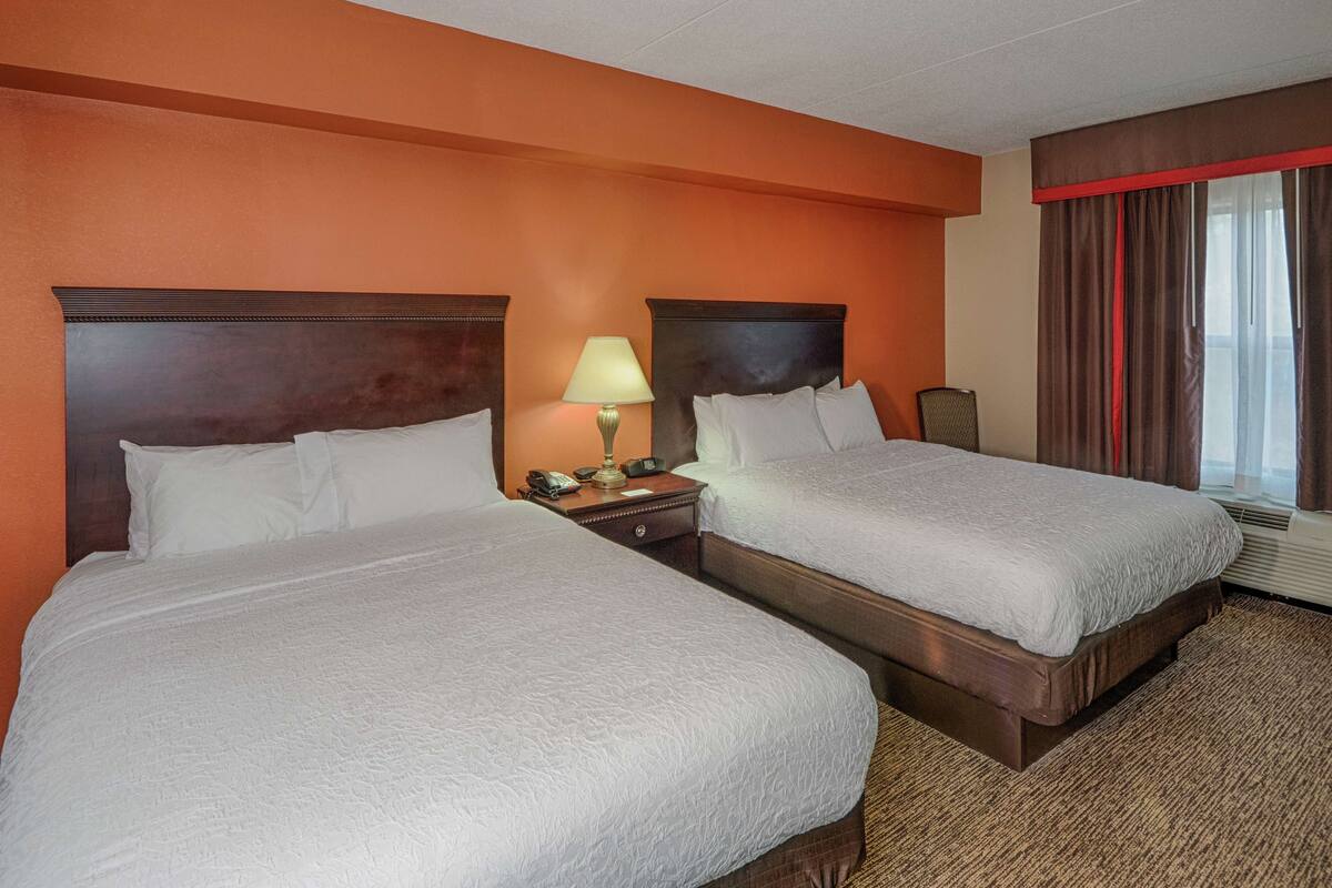 Ein Hotelzimmer mit zwei Betten, einem Nachttisch mit Lampe, einem Telefon und einem Fenster mit Vorhängen.