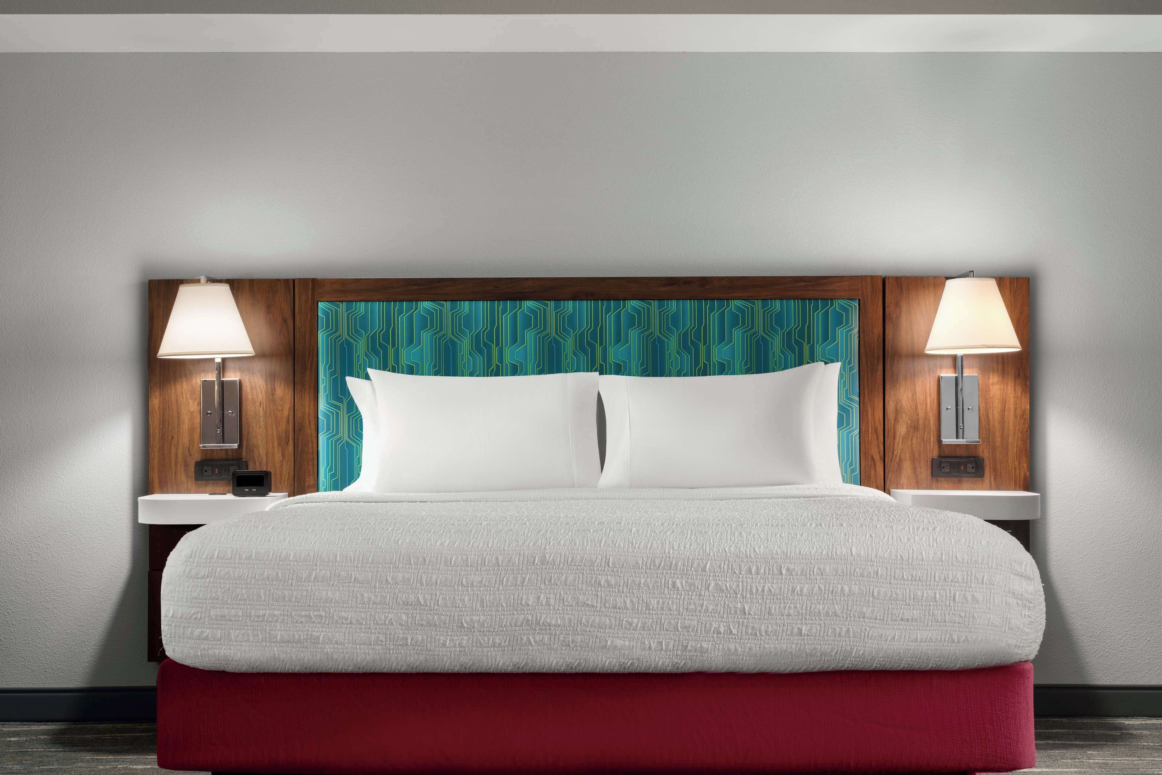 A neatly made bed with a red base, a white comforter, and two pillows. There are two bedside tables with lamps, and a wooden headboard with a teal geometric pattern.