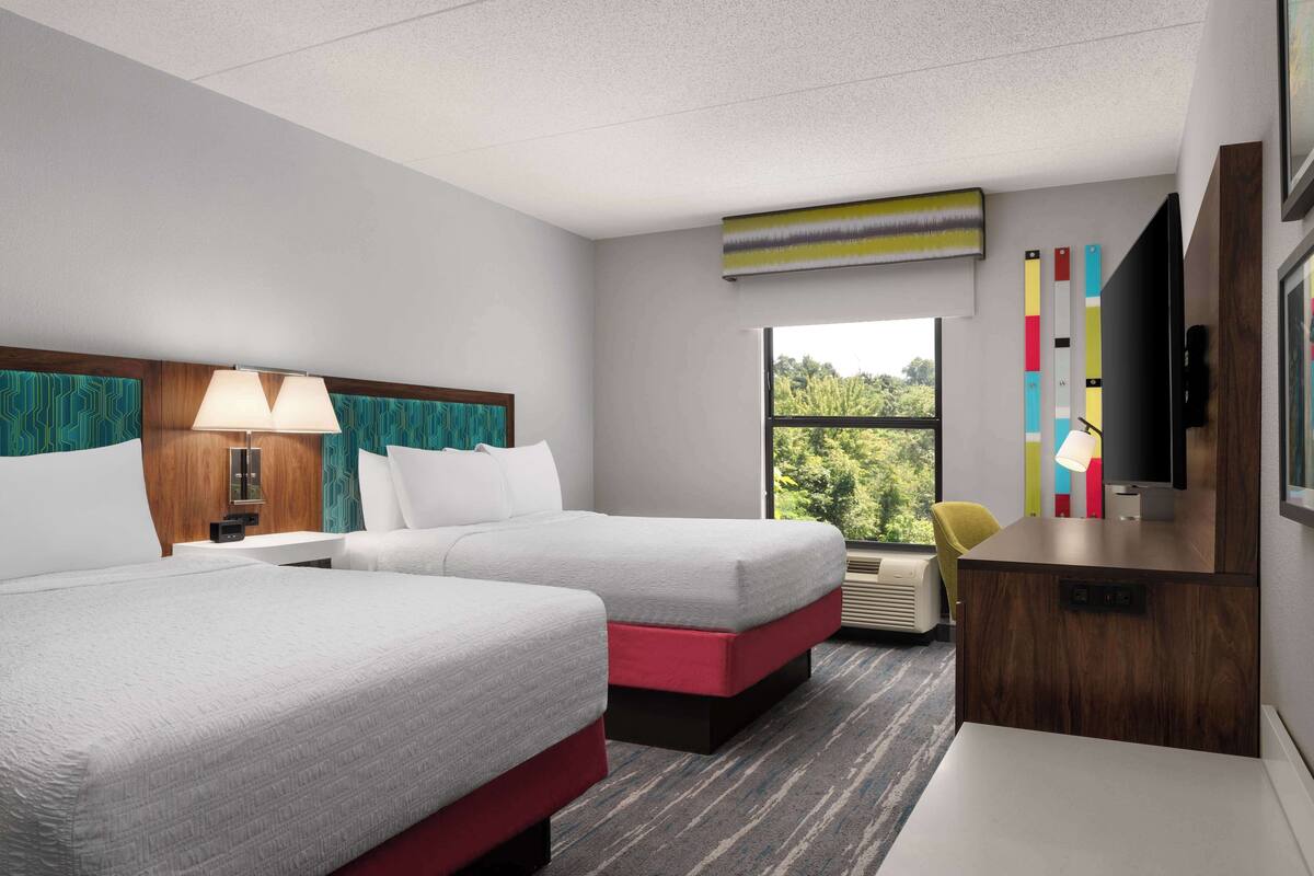 Ein Hotelzimmer mit zwei Betten, einem Schreibtisch, einem Fernseher und einem Fenster mit Blick auf Bäume.