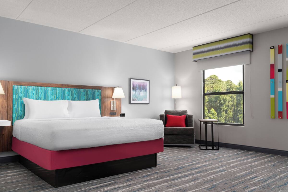 Ein Hotelzimmer mit einem Bett, einem Schreibtisch, einem Stuhl, einem Fenster und einem an der Wand hängenden Kunstwerk.