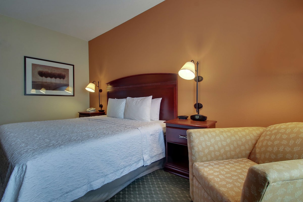 A hotel room with a bed, bedside lamps, a chair, a nightstand, and a framed picture on the wall.