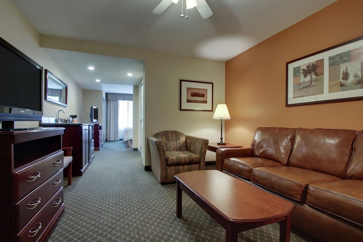A hotel room with a leather sofa, a wooden coffee table, a TV, a desk with drawers, a chair, and a lamp.
