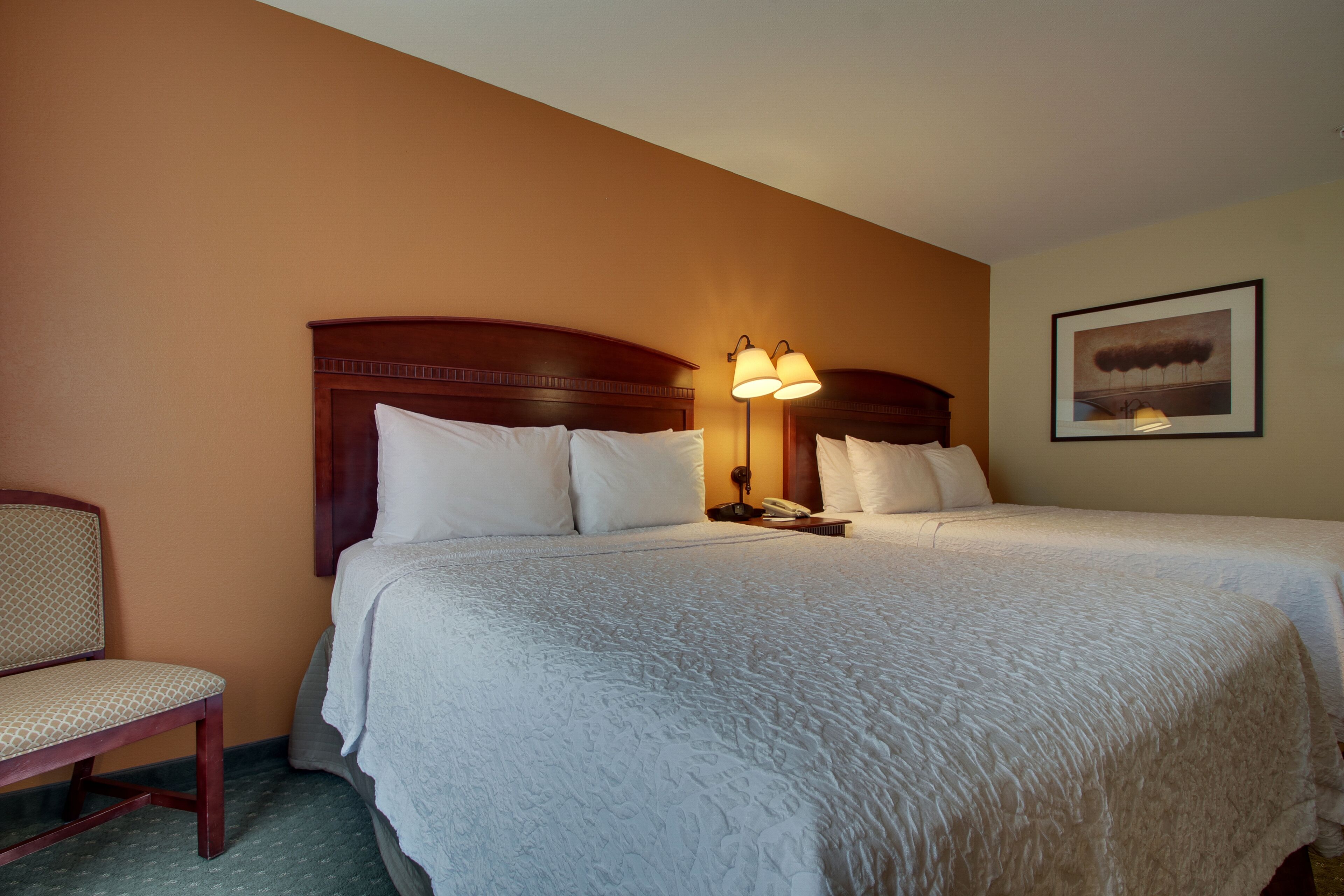 A hotel room with a large bed, a bedside table with a lamp, a chair, and a framed picture on the wall.