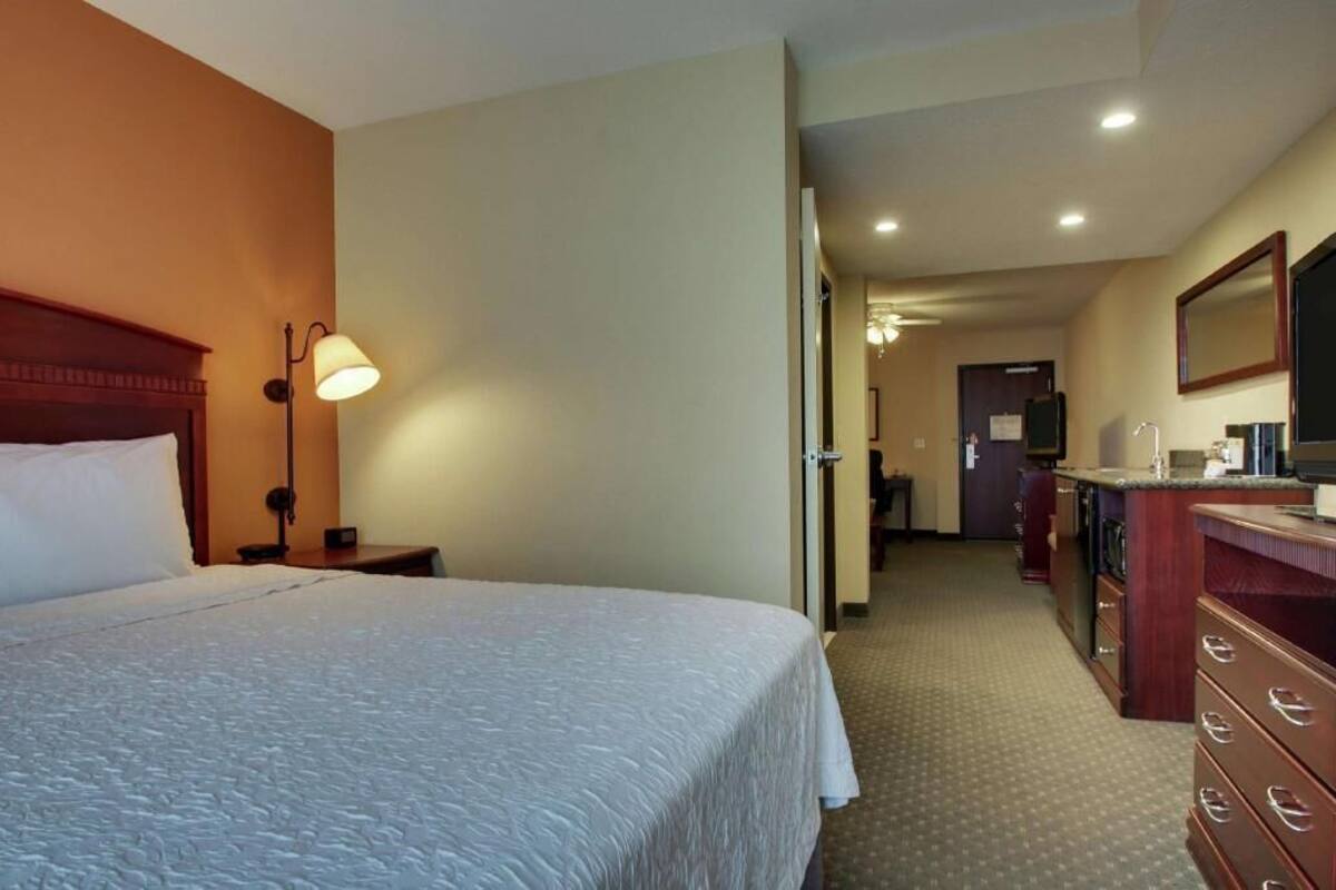 A hotel room with a large bed, a desk with a lamp, a dresser, and a mirror.