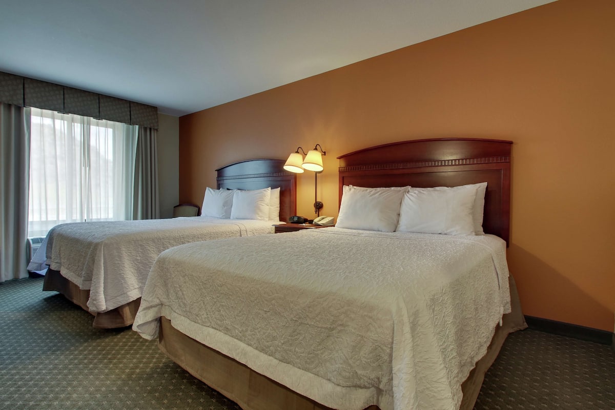 A hotel room with two beds, a nightstand with a lamp, and a window with curtains.