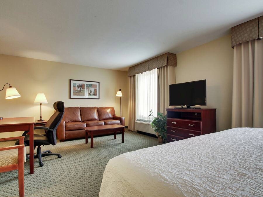 Ein Hotelzimmer mit einem Bett, einem Schreibtisch mit Stuhl, einer Ledersofa, einem Fernseher auf einer Kommode und einem gerahmten Bild an der Wand.