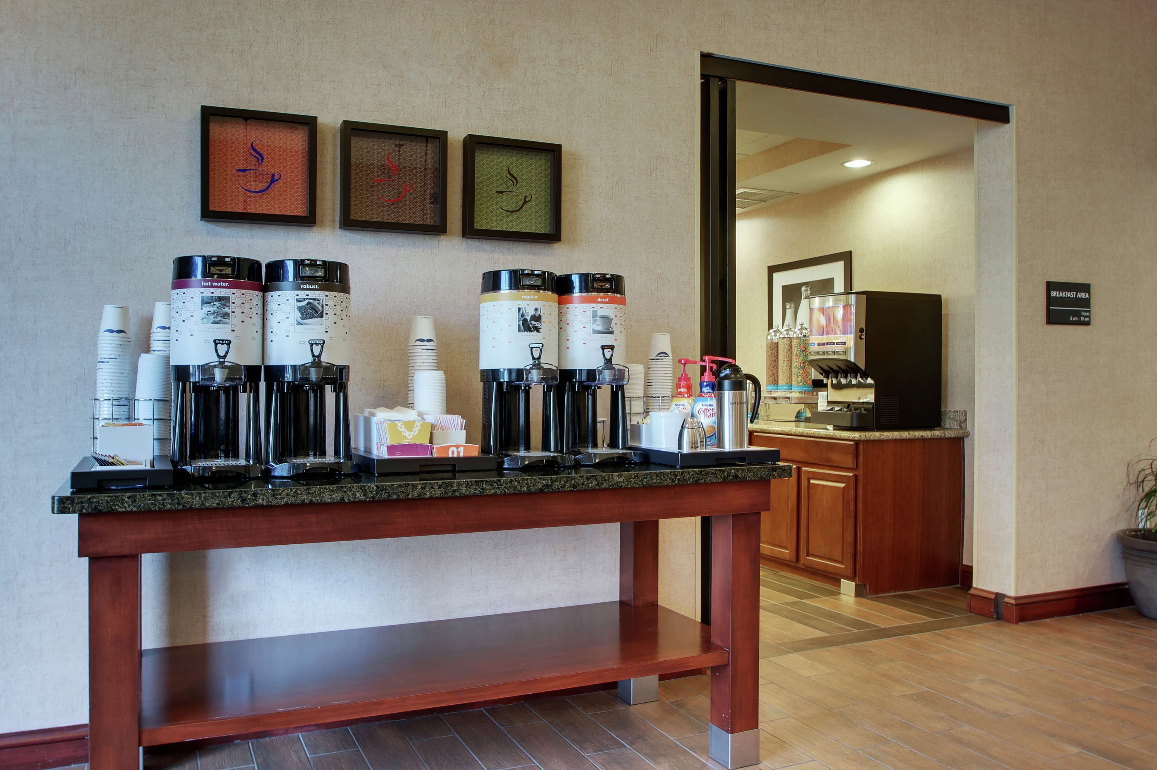 Un área de desayuno de hotel con varias máquinas de café, vasos desechables y un letrero que indica el espacio del desayuno.
