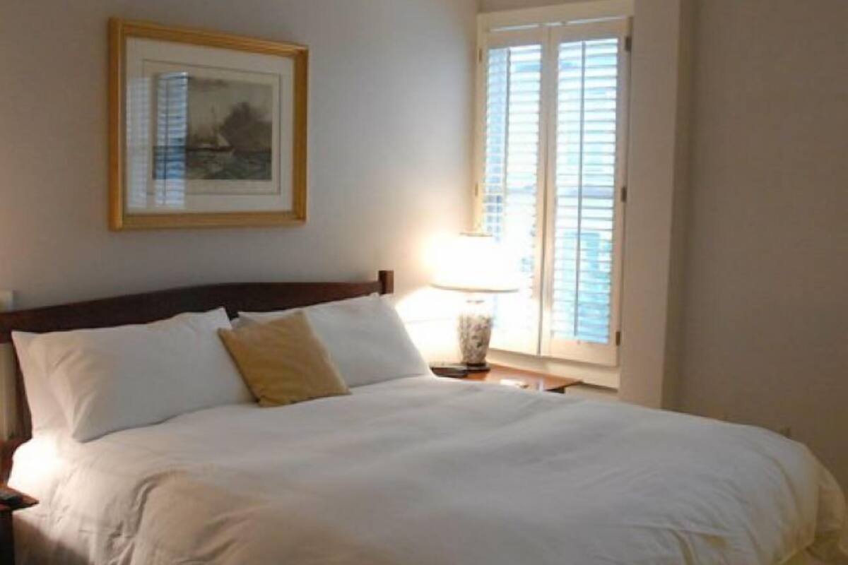 A bedroom with a large bed, a bedside table with a lamp, a framed picture on the wall, and a window with blinds.