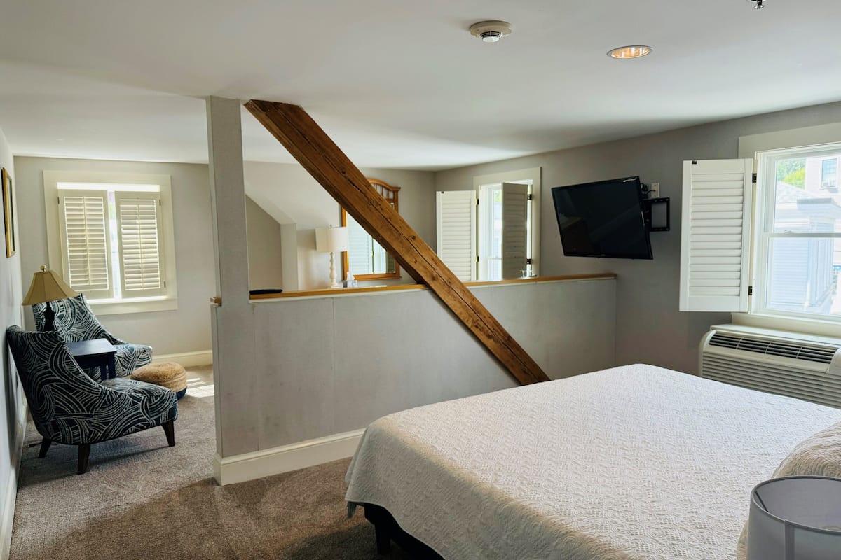 A bedroom with a large bed, a chair, a desk, a TV, and a window with shutters.