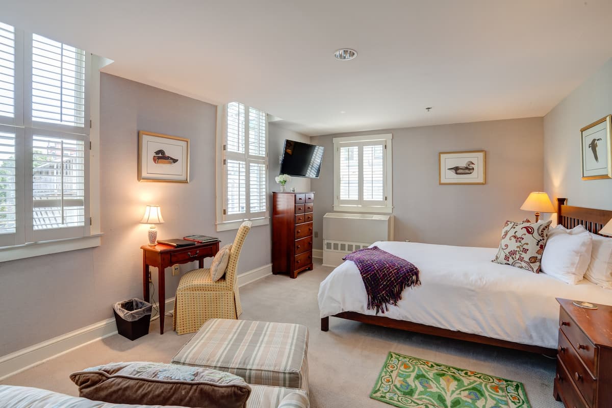 A bedroom with a bed, a desk, a chair, a TV, and a window.