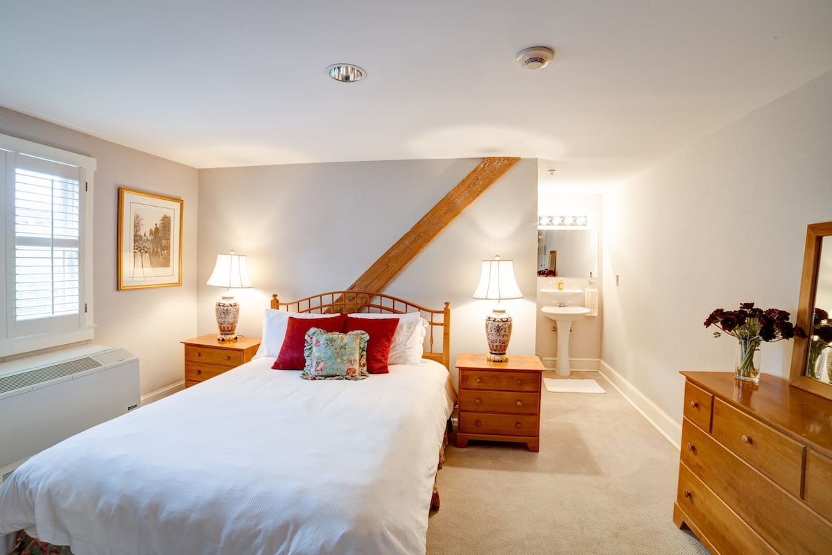 A bedroom with a large bed, two bedside tables with lamps, a wooden headboard, a window with blinds, a radiator, and a dresser with a mirror.