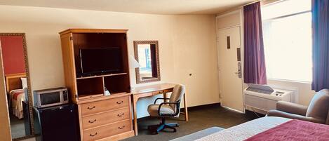 A hotel room with a bed, a desk with a chair, a wardrobe, a television, and an air conditioning unit.