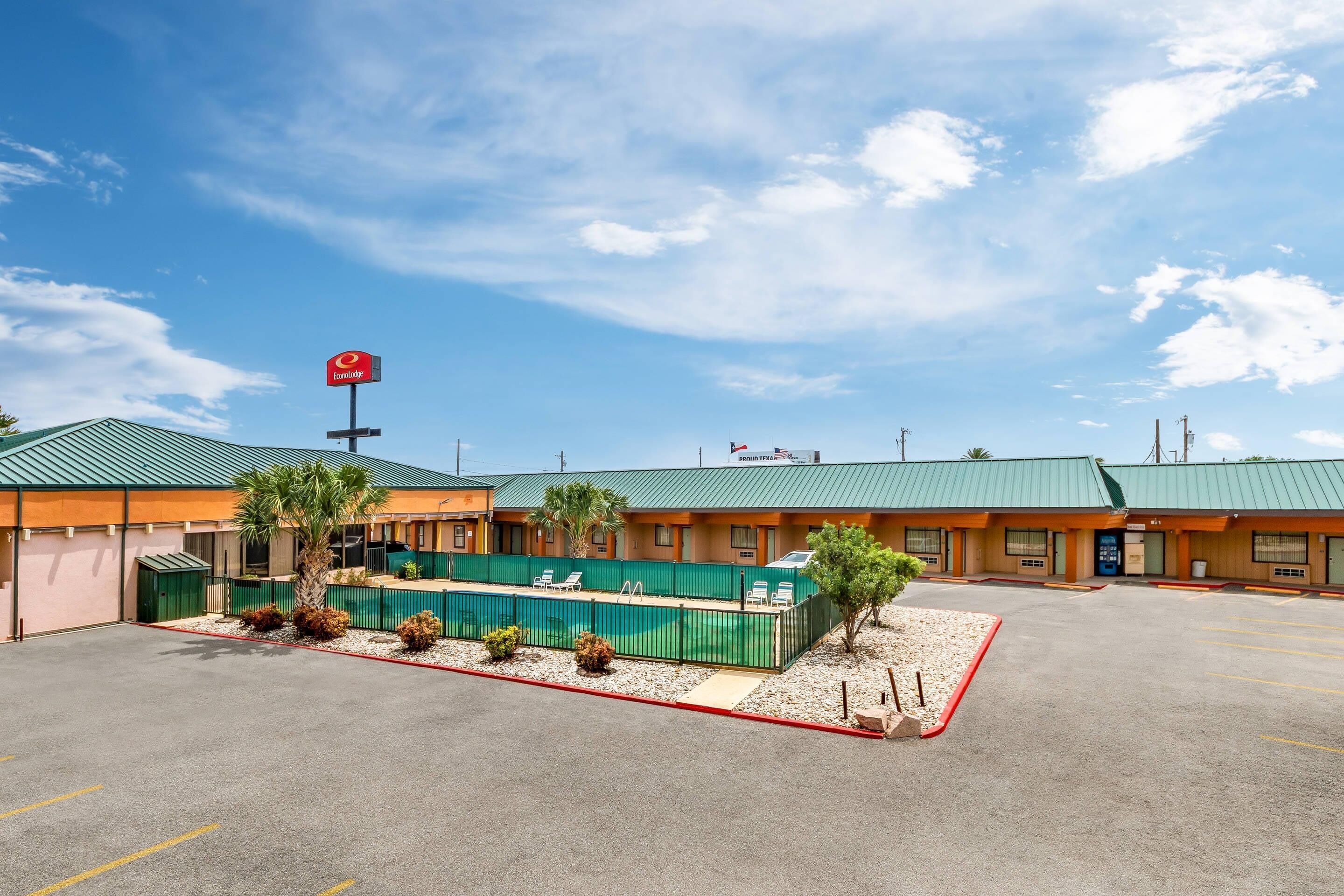 A motel with a green roof, a parking lot, and a fenced area with palm trees.