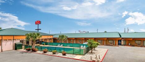A motel with a green roof, a parking lot, and a fenced area with palm trees.