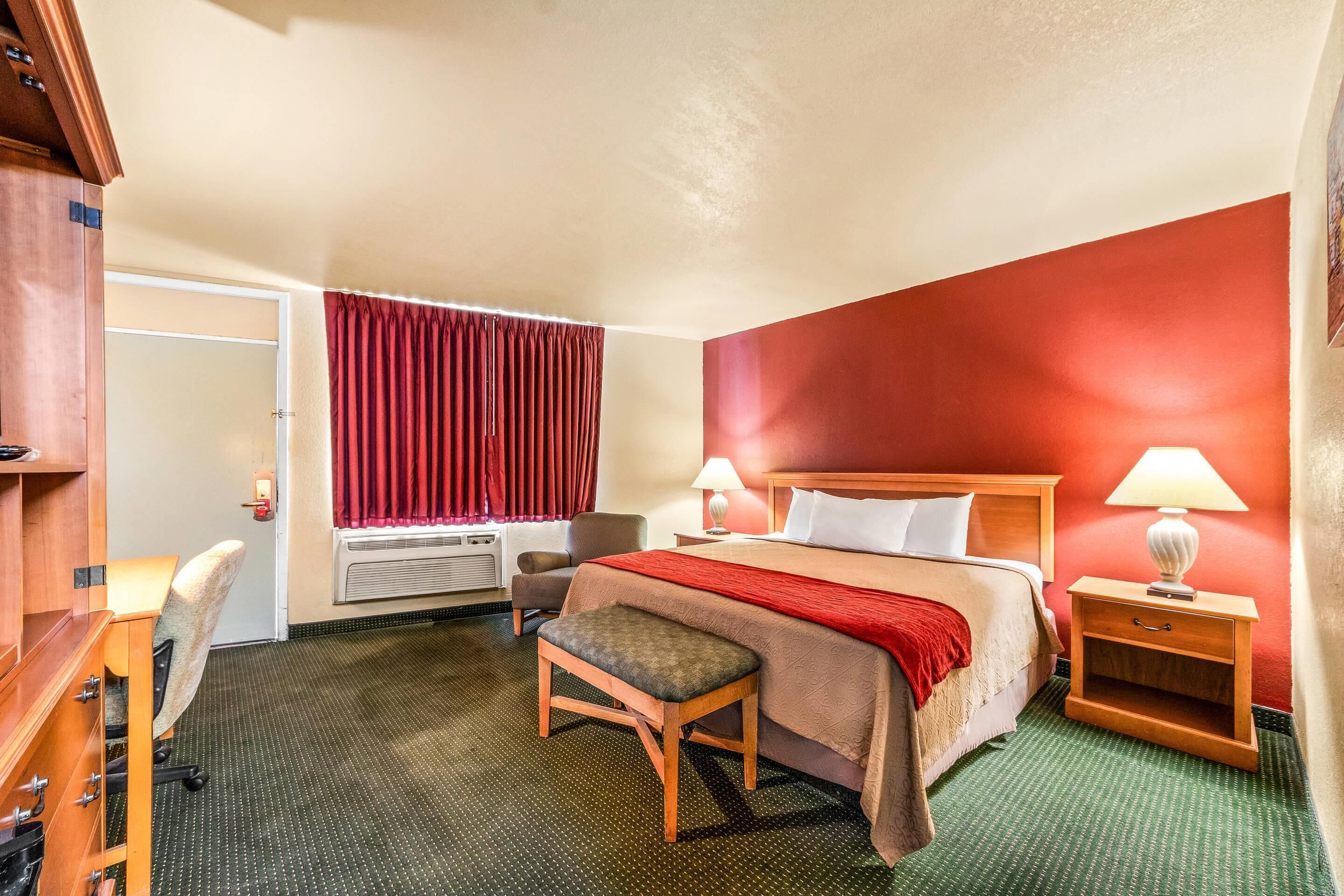 Een hotelkamer met een bed, nachtkastje, lamp, stoel, bureau en raam met gordijnen.