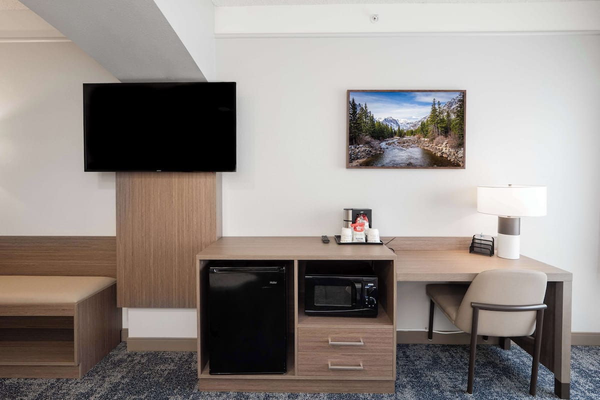 Une chambre d’hôtel équipée d’un téléviseur à écran plat fixé au mur, d’un bureau avec un micro-ondes, d’un réfrigérateur, d’une chaise et d’une petite table avec une lampe.