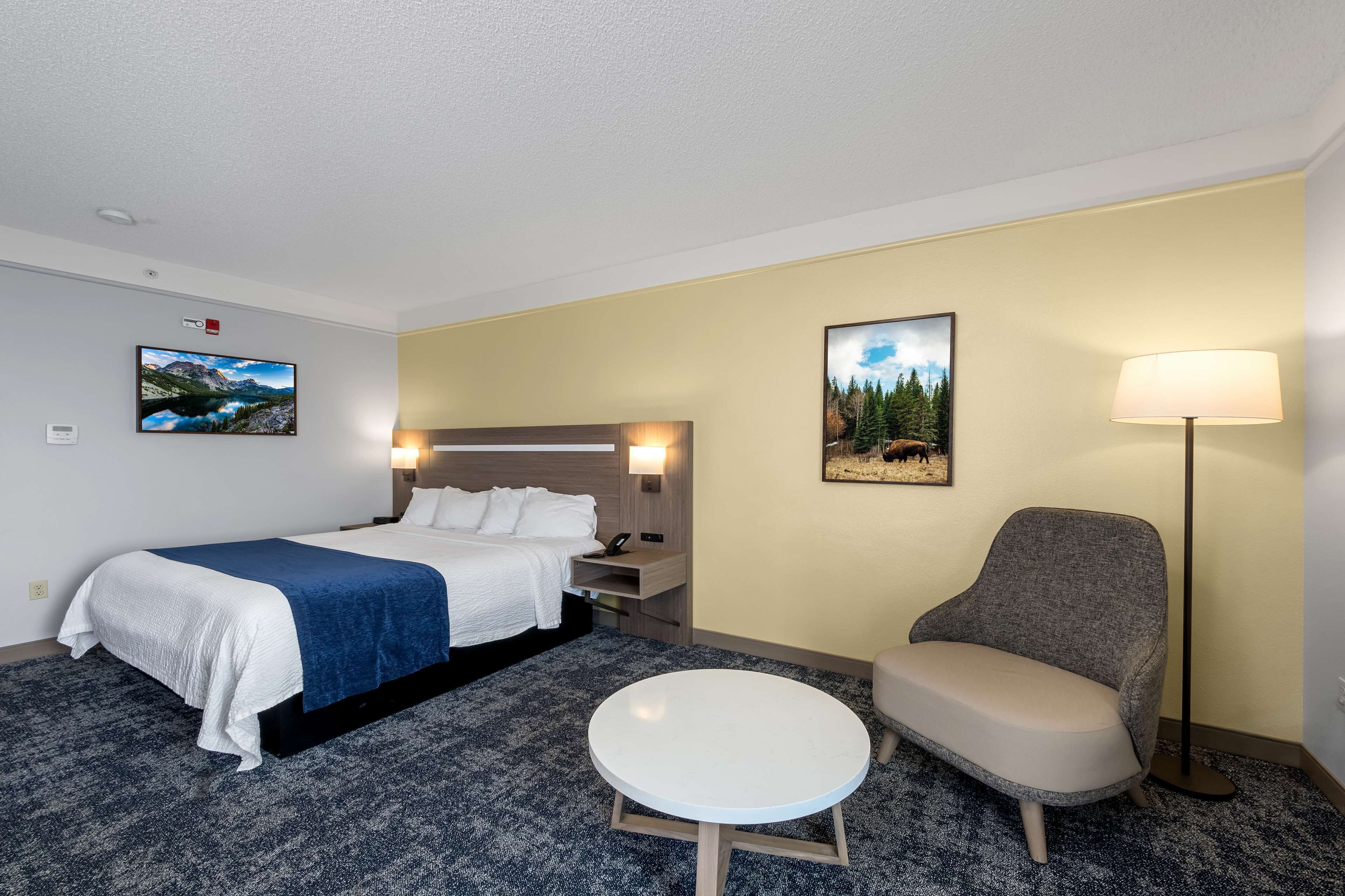A hotel room with a bed, a round table, a chair, a lamp, and two wall-mounted pictures.