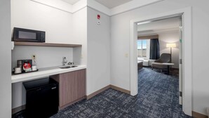 Une chambre d’hôtel compacte dotée d’une kitchenette, d’un four à micro-ondes, d’un évier et d’un lave-vaisselle. Une porte donne accès à un coin salon avec une chaise et une lampe.