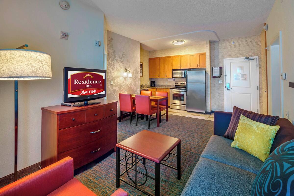 A hotel room with a sofa, a small table, a TV, a lamp, a kitchenette, and a dining area.