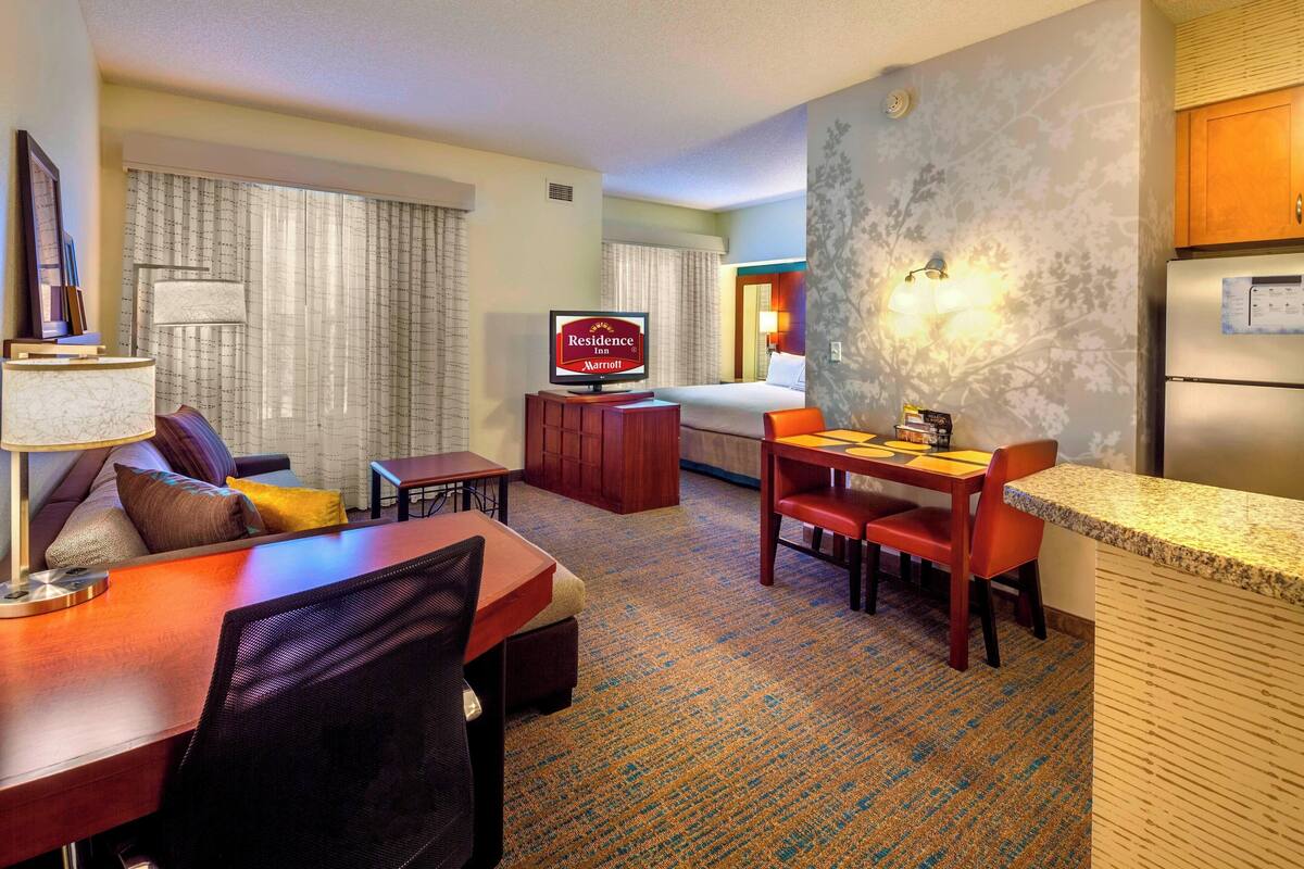 A hotel room with a bed, a TV, a desk with a chair, a small table, and a refrigerator.