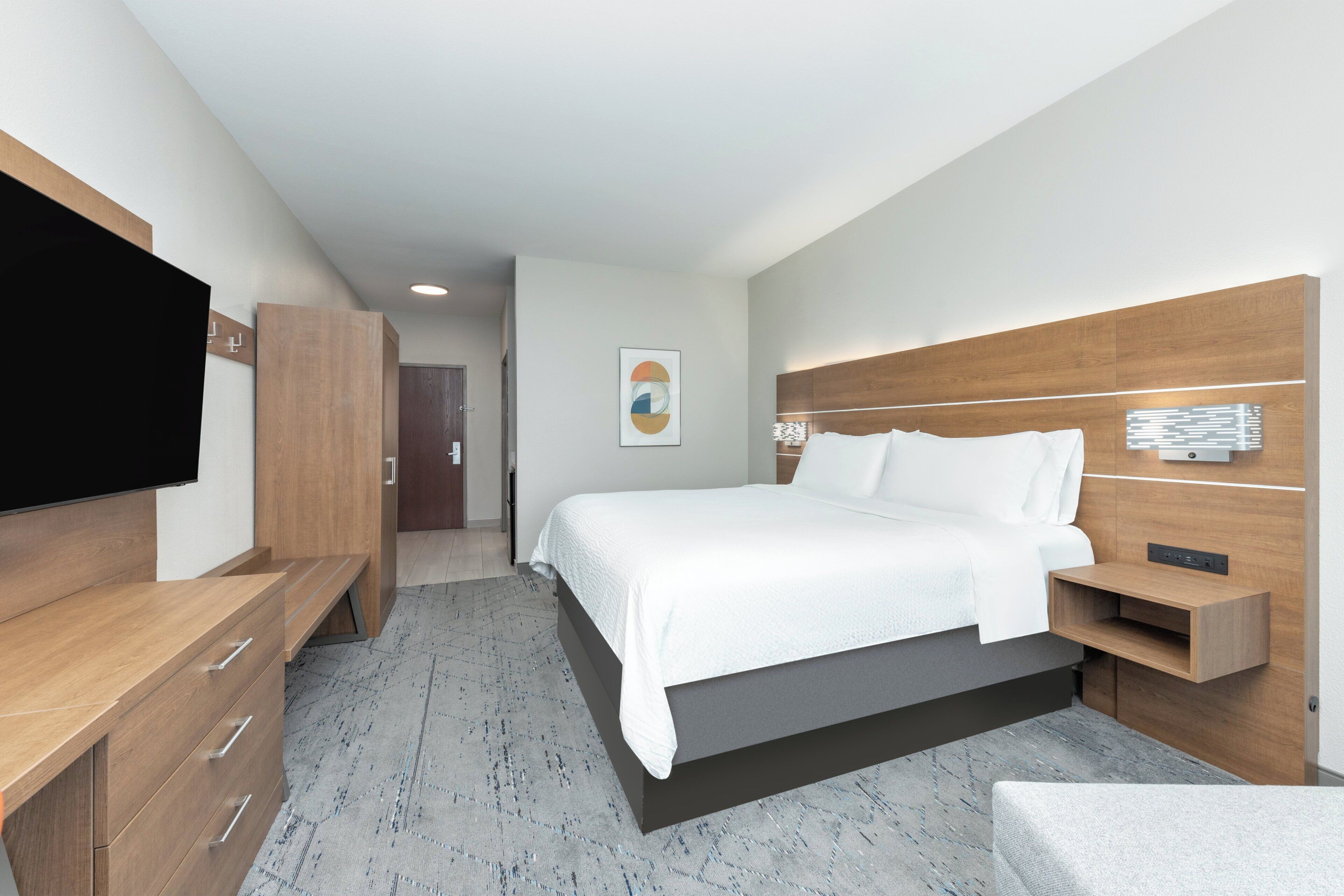 A modern hotel room with a large bed, wooden furniture, a flat-screen TV, and a wall-mounted light fixture.
